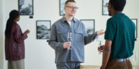 Group of diverse people mingling and discussing art in gallery space featuring framed artworks. Guests holding wine glasses and engaging in conversation about exhibits