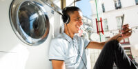 Portrait young arab man sitting against washing machine listening to music with phone and headphones